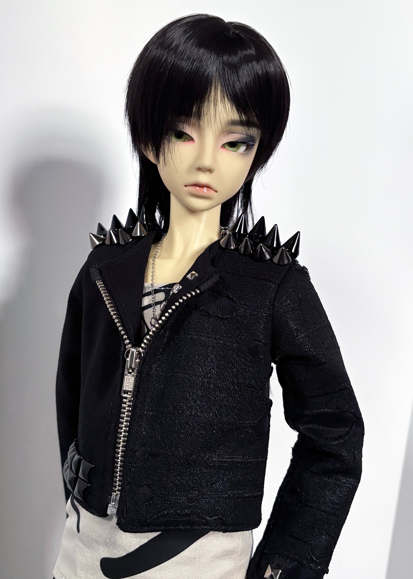A doll with short black hair wearing a black textured zip-front jacket with spiked shoulders, paired with a patterned shirt and light bottoms, photographed against a plain light background.