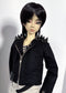 A doll with short black hair wearing a black textured zip-front jacket with spiked shoulders, paired with a patterned shirt and light bottoms, photographed against a plain light background.