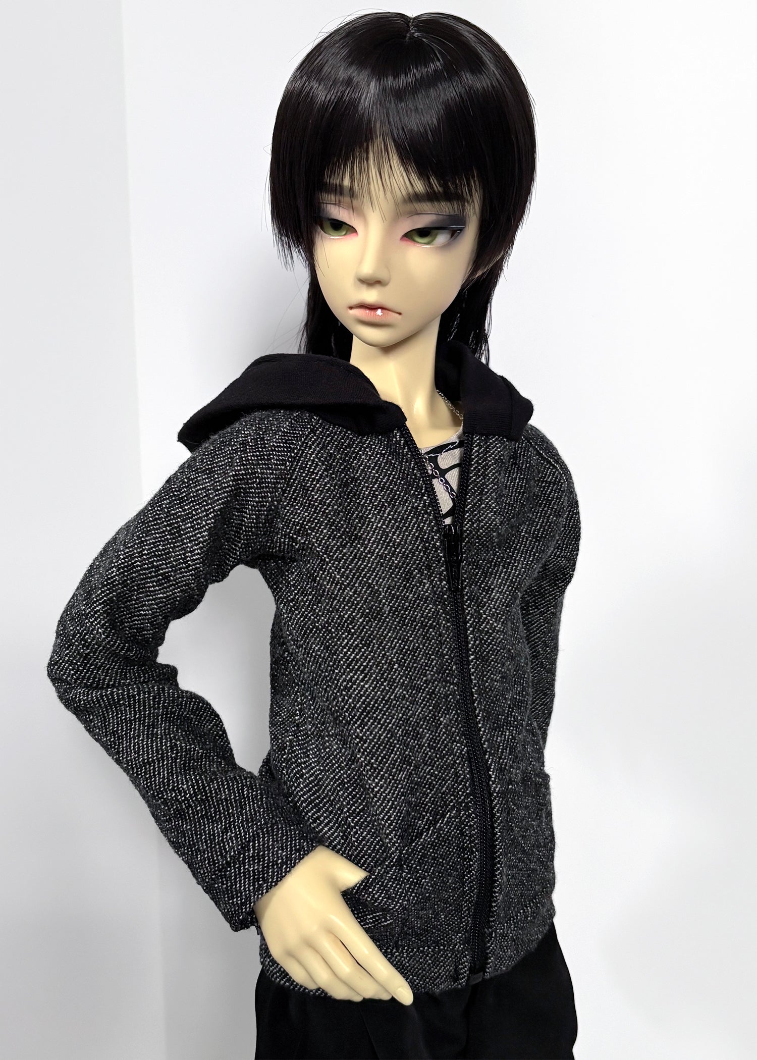 A doll with short black hair wearing a dark gray and black textured zip-up jacket with a black hood, posed against a plain light background.