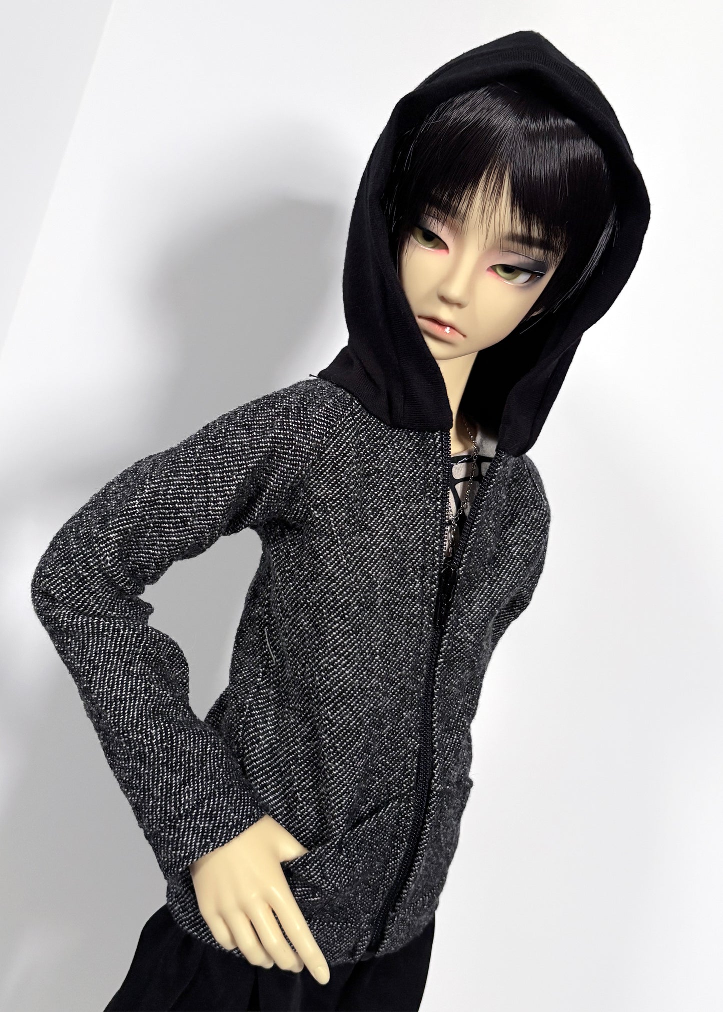 A doll wearing a black hooded zip-up jacket with a textured gray and black wool body, posed against a plain light background.