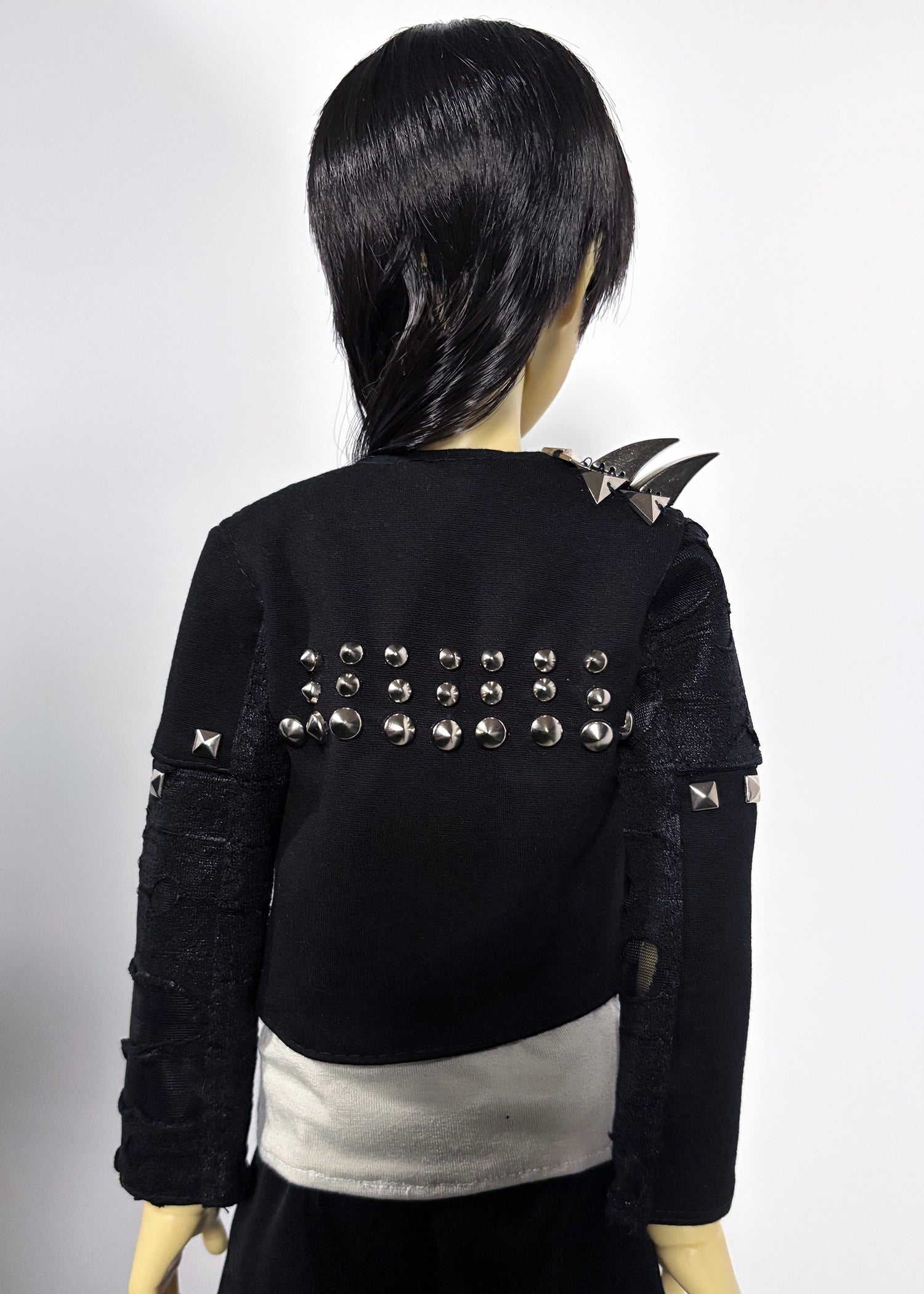 Back view of a black cropped jacket with silver studs and shoulder spikes, worn by a doll with black hair against a light gray background.