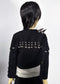 Back view of a black cropped jacket with silver studs and shoulder spikes, worn by a doll with black hair against a light gray background.