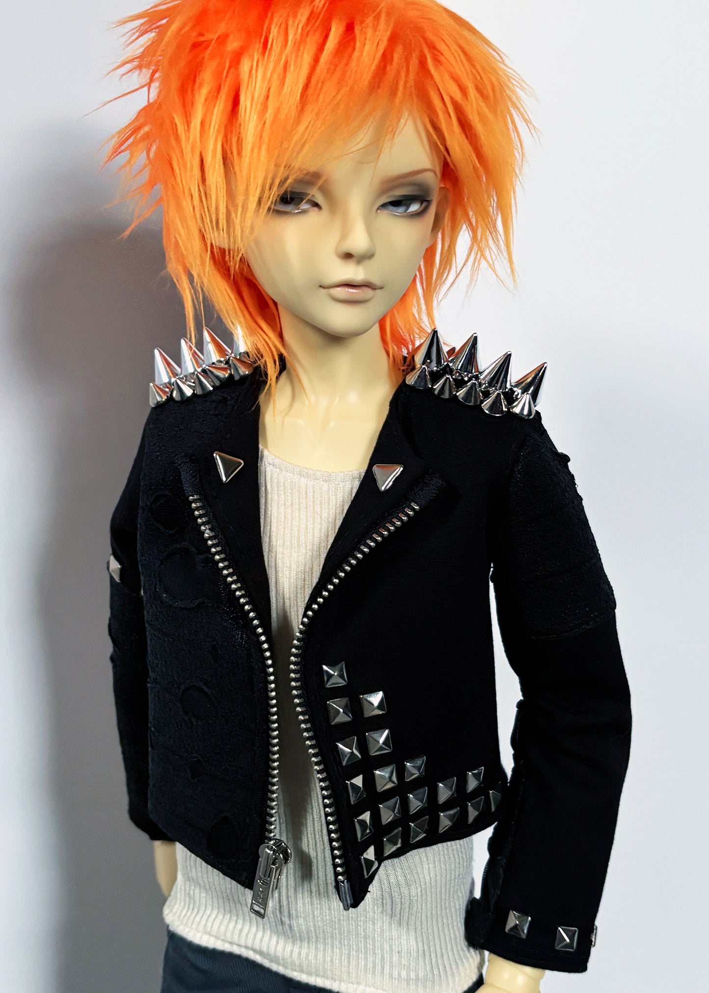 A doll with bright orange hair wearing a black studded moto jacket over a white ribbed top, photographed against a plain light gray background.