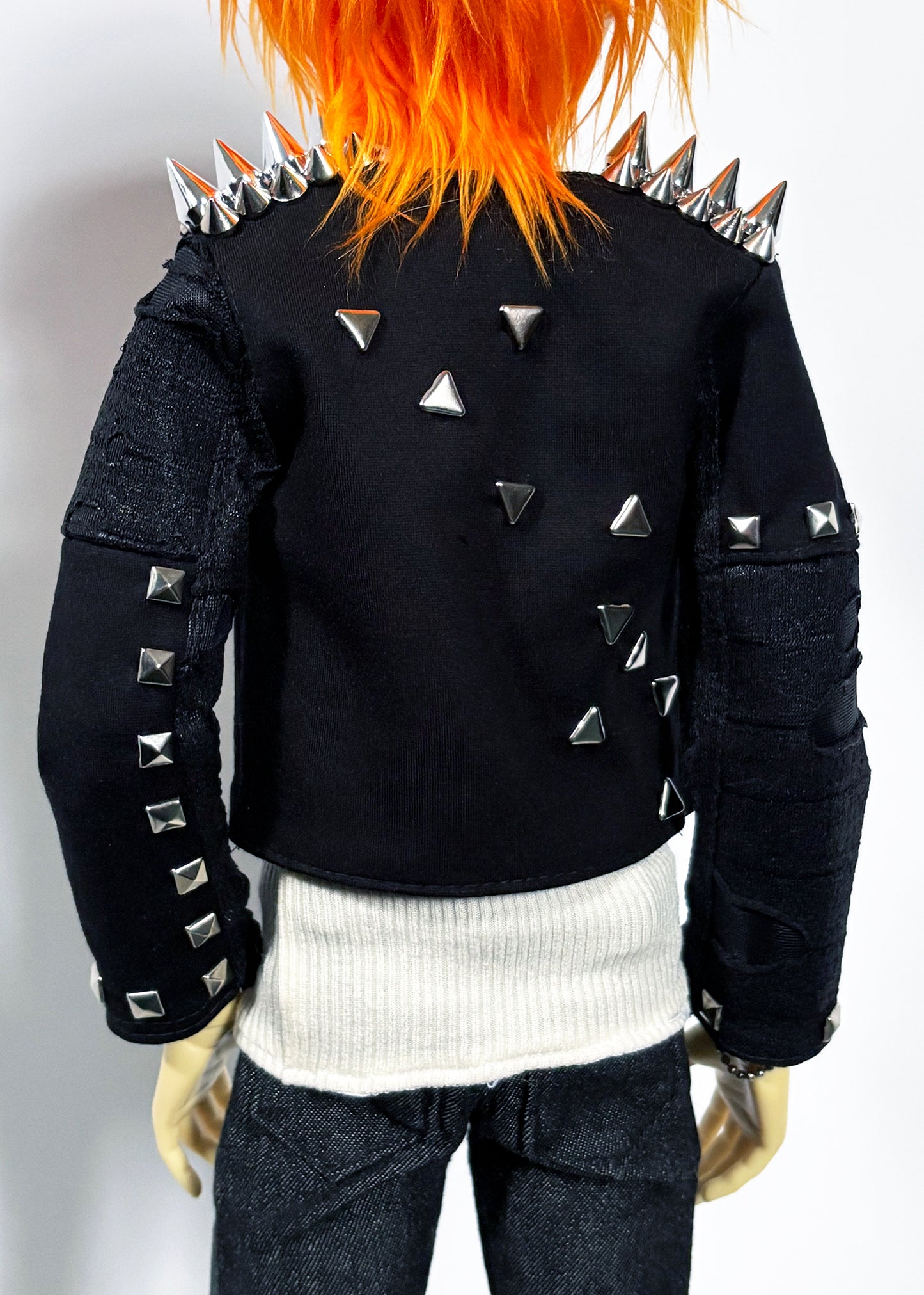A black studded jacket with silver metal spikes on the shoulders and sleeves, worn by a doll with bright orange hair, a white ribbed shirt, and dark jeans against a plain white background.