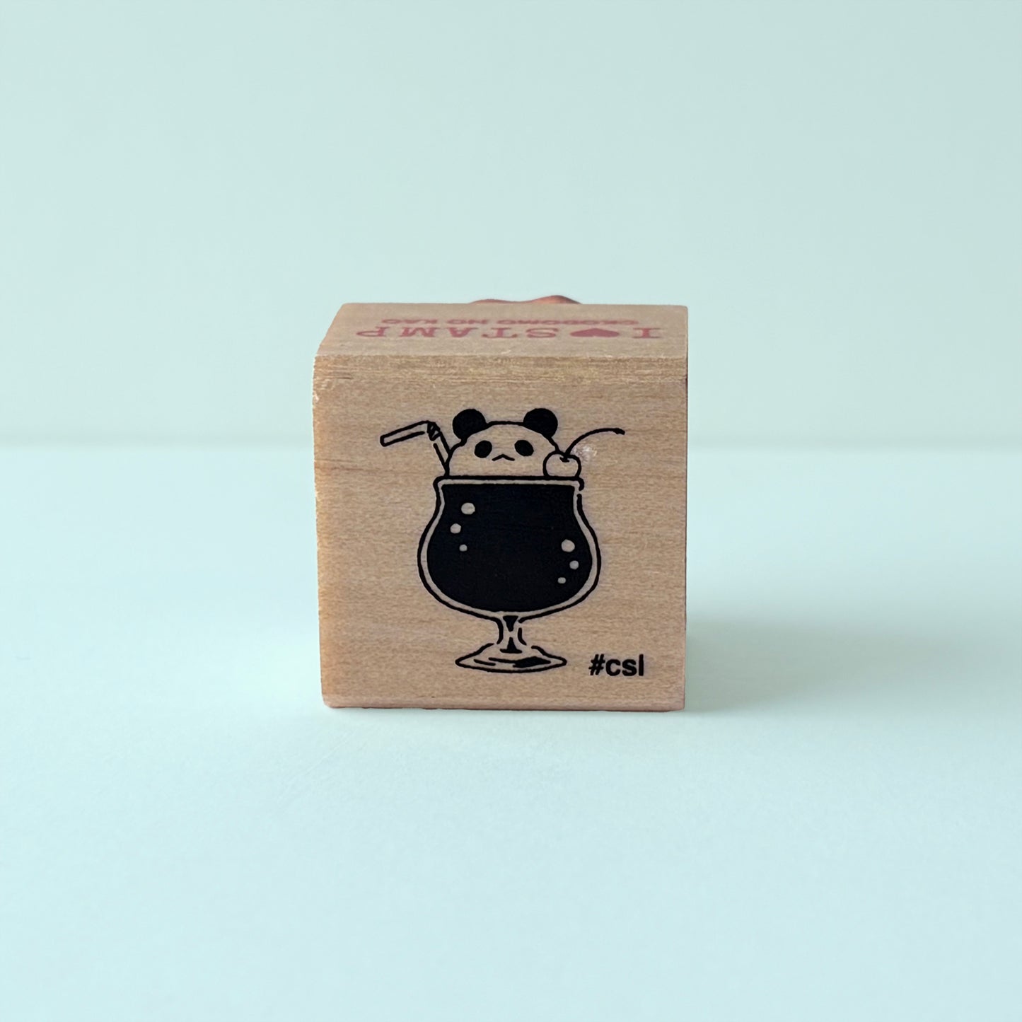 Wooden block stamp with a black illustration of a panda bear in a glass of soda on a light blue background