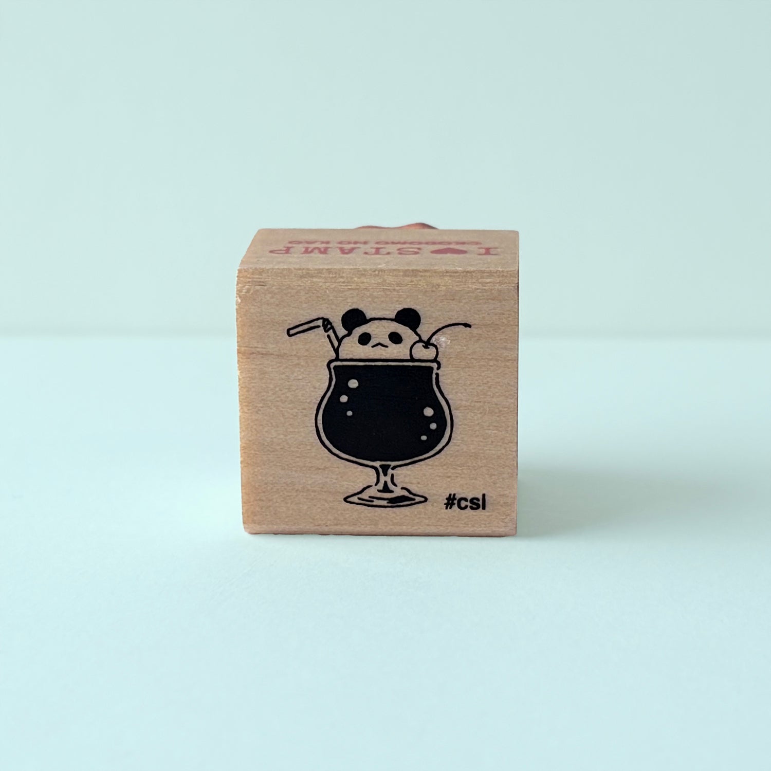Wooden block stamp with a black illustration of a panda bear in a glass of soda on a light blue background