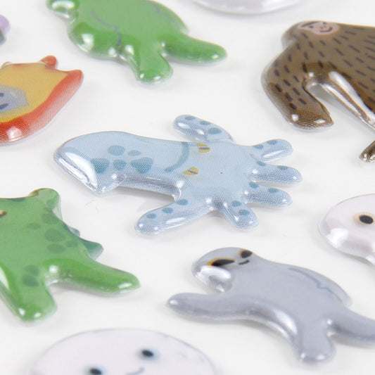 A close-up of glossy, animal-shaped stickers scattered on a white background, including a light blue octopus, a green dinosaur, a brown spotted creature, and others partially visible.